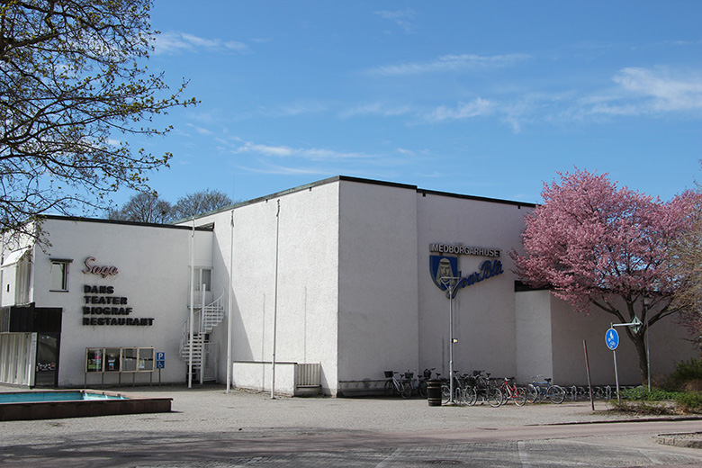Foto på medborgarhuset Joar Blå som det ser ut idag. Det är en vit stenbyggnad. Framför byggnaden i bilden syns en del av den större fontänen som ligger där. Man ser också cykelställ och blommande körsbärsträd.
